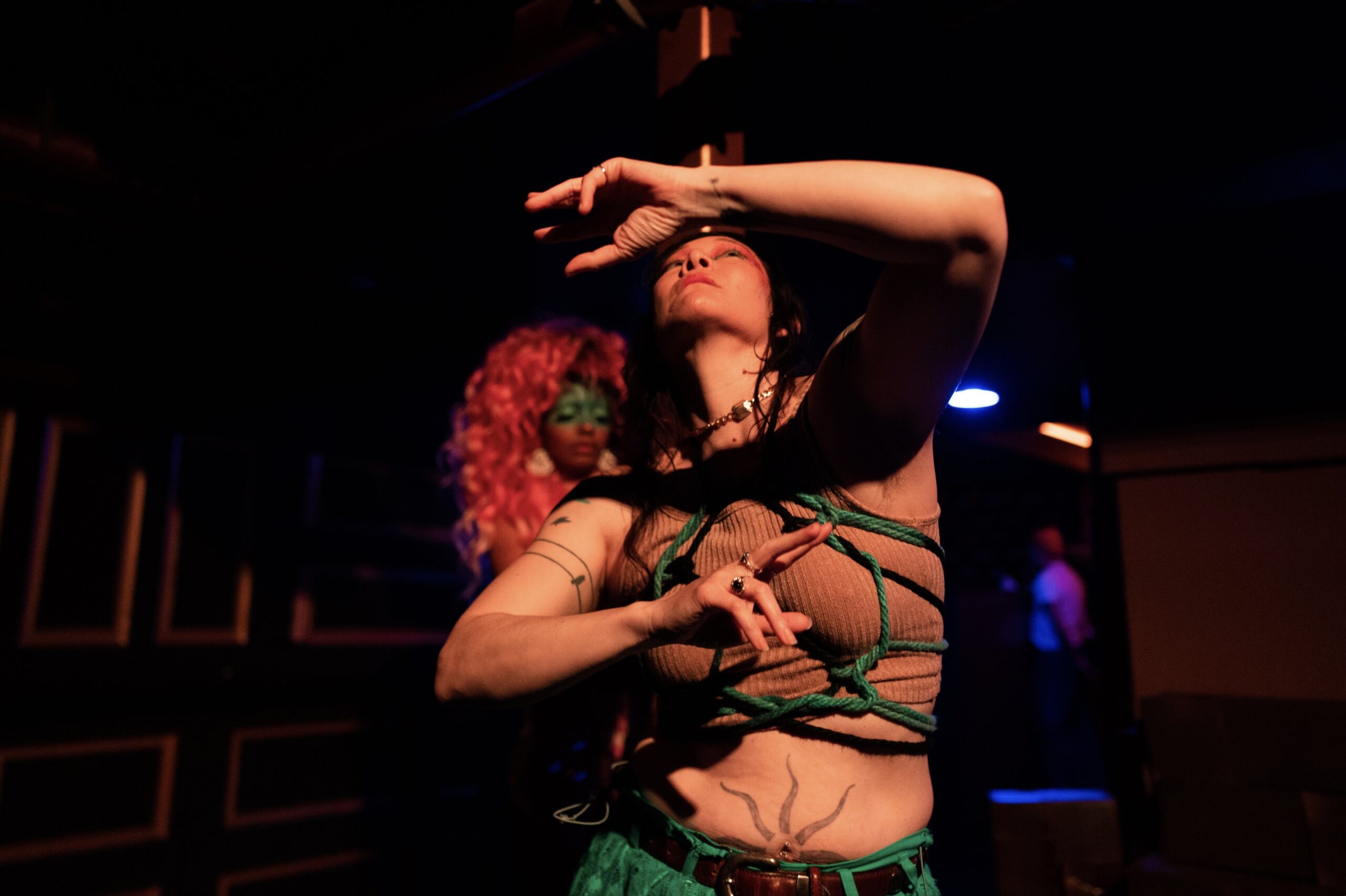 Erin Mei-Ling Stuart, a performer with teal shibari ropes wrapped around their torso, strikes an expressive pose under warm, dramatic lighting, while another artist with vibrant pink curly hair and green face paint stands in the background, adding to the atmospheric scene.